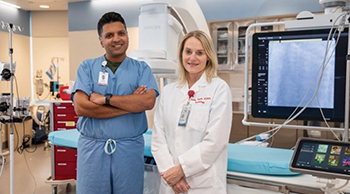 Male physician with short black hair, in blue scrubs with badge, arms crossed and female APRN, with blond shoulder-length hair and a white lab coat, posing in front of medical equipment and monitors.