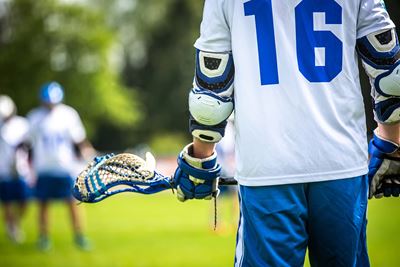 Lacrosse player holding an empty crosse, with a white and blue jersey makred "16"and blue shorts, on a green field with blurred players and trees in the background. The player is pictured from shoulder to upper thigh.