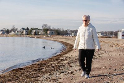  older woman with white hair and white jacket, dark pants and sunglasses, walking on calm sandy beach with buildings and trees in background on horizon - mending broken hearts