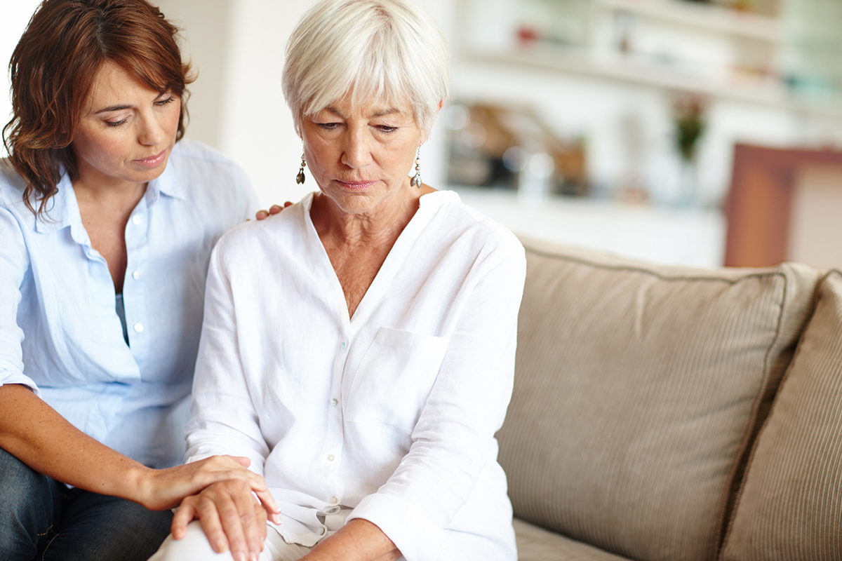 An older woman with white hair, wearing a white top, sits on a couch, being comforted by a younger woman in a blue top. The younger woman has her hand on the older woman's hand.
