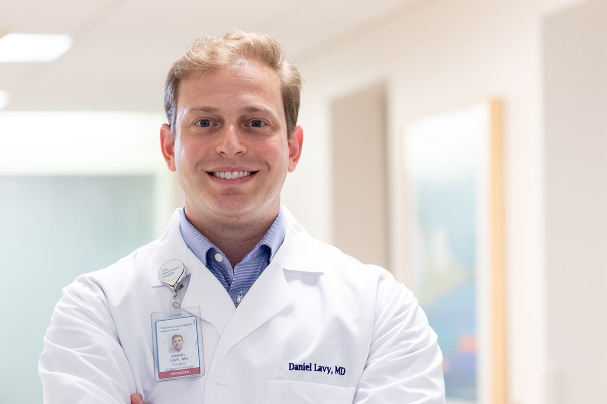 A smiling, young, white man with blond hair poses in a hospital hallway. He is wearing a white clinical jacket with an ID badge.