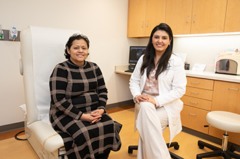 Two women pose, seated, in a hospital exam room, one in a  plaid outfit, the other in a white clinical jacket. Both are smiling.